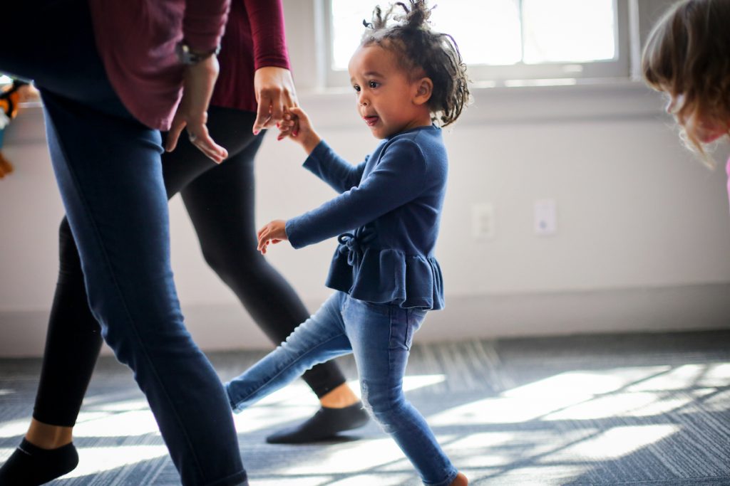 A 2-year-old girl dances across the room with her mother during a group music class for toddlers.