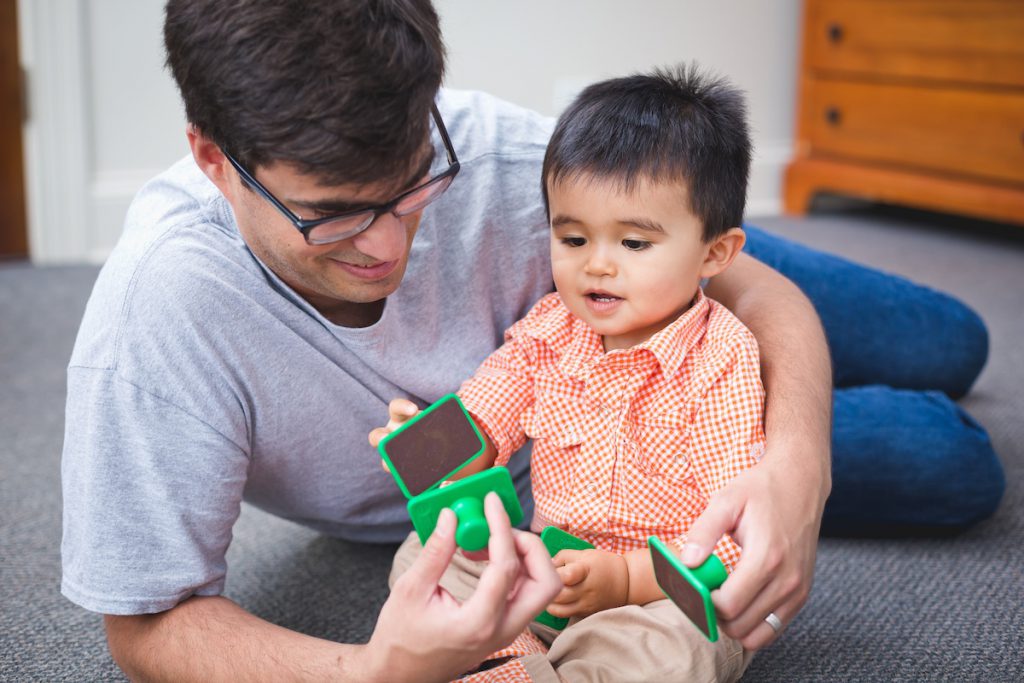 A dad taps sandblocks on the floor with his toddler son during a Kindermusik class—one of the easiest rhythmic early literacy strategies.